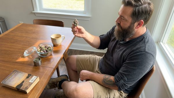 Man examining a frosty, colorful cannabis bud at a kitchen table with a bowl of buds and coffee mug.